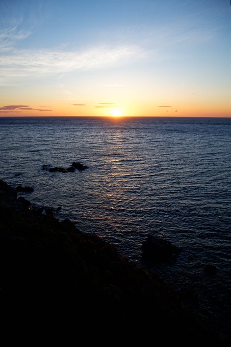 A sunset over the ocean with the sun dipping low on the horizon, casting a golden reflection across the water, while silhouetted rocks and a dark coastline frame the lower portion of the scene—evoking the beginning of night, aligning with the "GN" (good night) sentiment.