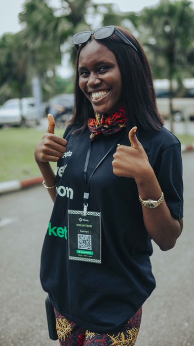 A smiling young woman giving two thumbs up, wearing sunglasses on her head, a black t-shirt with sponsor logos, and a volunteer lanyard with a QR badge.