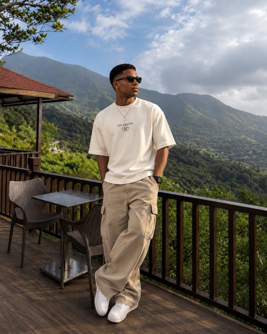 A young man wearing a cream "LES ANGELES" t-shirt, tan cargo pants, and white sneakers poses on a mountain-view balcony, embodying a calm, focused start to the day that reflects the post's message of making small, meaningful progress today.
