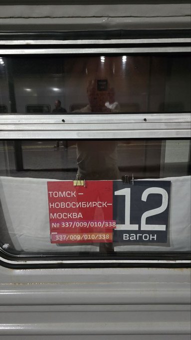 A train window displays a red and black sign indicating route 'ТОМСК – НОВОСИБИРСК – МОСКВА № 337/009/010/338' and car number '12 вагон', with the photographer's reflection visible.