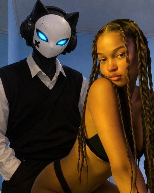 A photograph showing a person wearing a white cat-like mask with glowing blue eyes and black headphones, dressed in a black vest over a white shirt, posing next to a woman with long braided hair wearing black.
