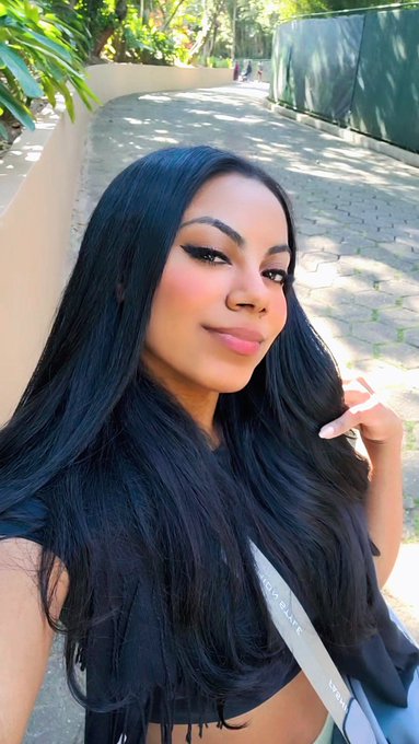 A selfie of a woman with long dark wavy hair, wearing makeup and a black top, standing outdoors in a sunlit courtyard with greenery.