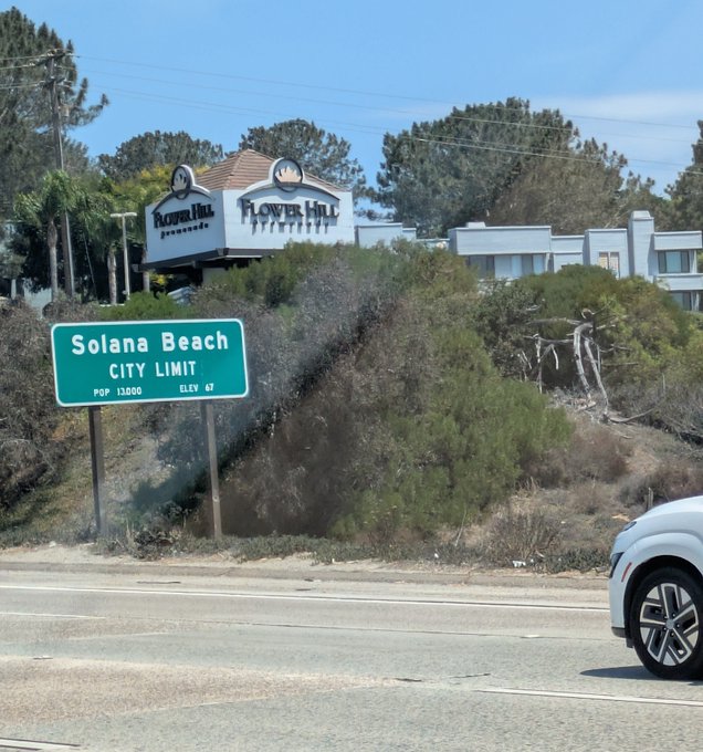 A green highway sign marks the "Solana Beach City Limit" (Pop. 13,000, Elev. 67 ft), visible just below a large "Flower Hill Promenade" sign, while a white car drives along the road into the California coastal city.