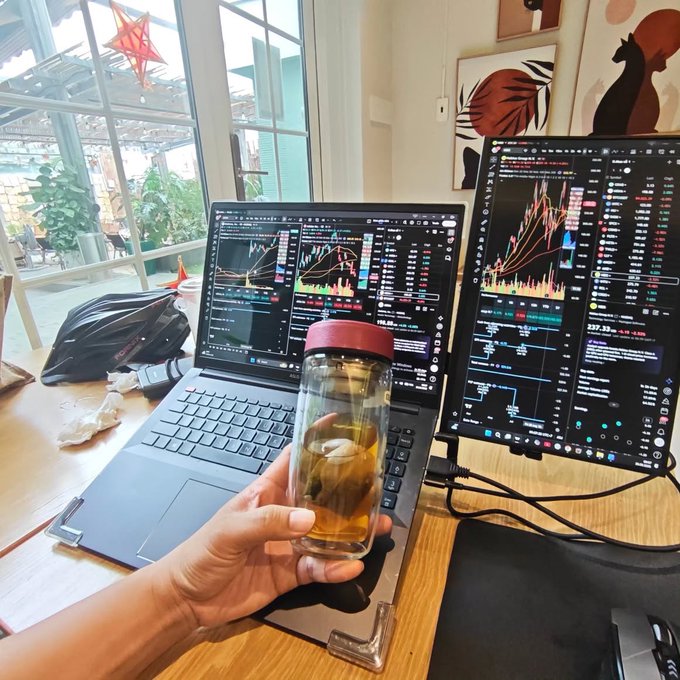 A person's hand holding a glass infuser bottle of tea in front of a dual-monitor trading setup displaying financial charts.