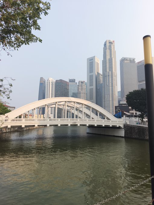 A daytime photograph of a white arched bridge spanning a river, with a backdrop of modern skyscrapers and high-rise structures in a hazy cityscape.