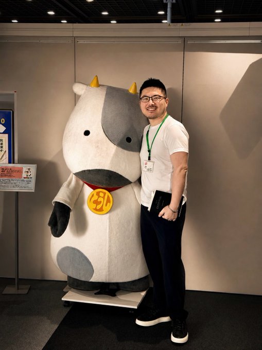 A man wearing glasses and a green lanyard poses proudly next to a large plush cow mascot with horns and a bell, holding what appears to be a laptop or notebook. The playful caption frames the cow-themed mascot as a "proof of patience," symbolizing the bullish optimism of holding through market ups and downs while aspiring toward a $200K milestone.