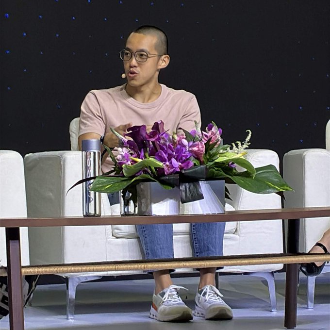 A man with glasses and a pink t-shirt sits on a white chair at what appears to be a panel or talk show, with a floral arrangement of purple orchids on the coffee table in front of him.
