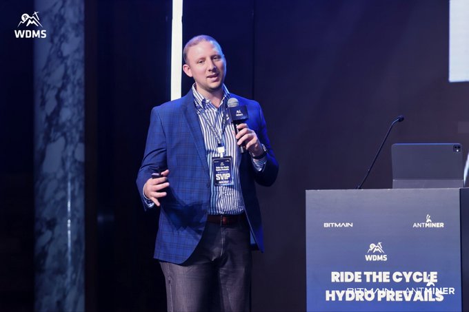 A man in a blue plaid blazer speaking into a microphone at a podium labeled "RIDE THE CYCLE HYDRO PREVAILS" with Bitmain, Antminer, and WDMS logos.