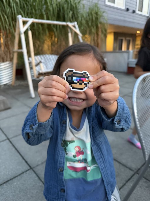A young child holding up a pixel art sunglasses sticker in front of their eyes, smiling and standing outdoors on a patio with chairs and plants visible.