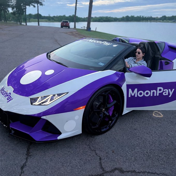 A person sits in a purple and white Lamborghini convertible decorated with MoonPay branding, parked near a lake.