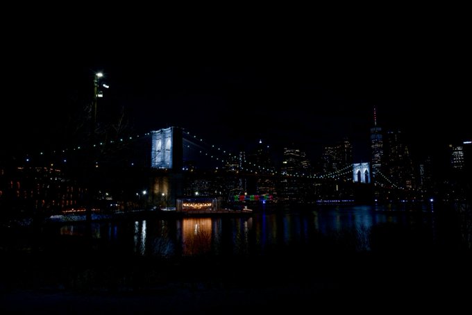 A nighttime view of the Brooklyn Bridge illuminated over the East River, with the Manhattan skyline visible in the background.
