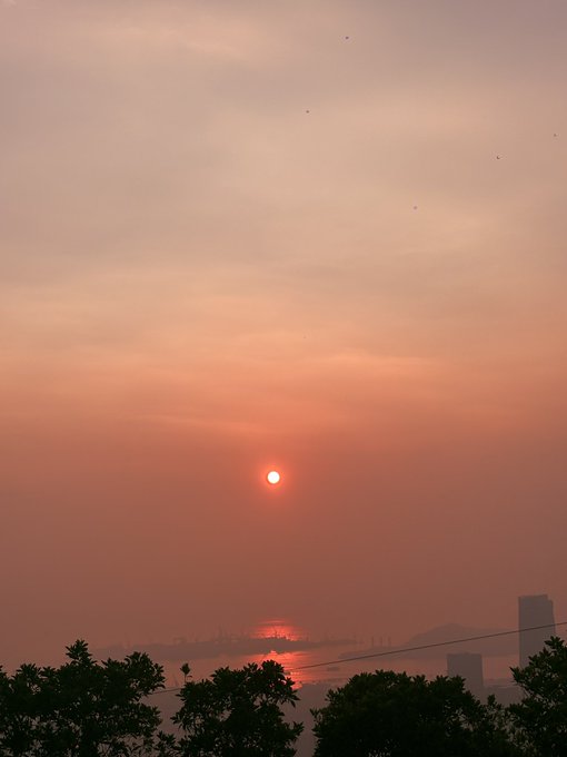 A serene sunset photograph showing a reddish-orange sun over a hazy cityscape with water in the foreground and silhouetted trees at the bottom edge.
