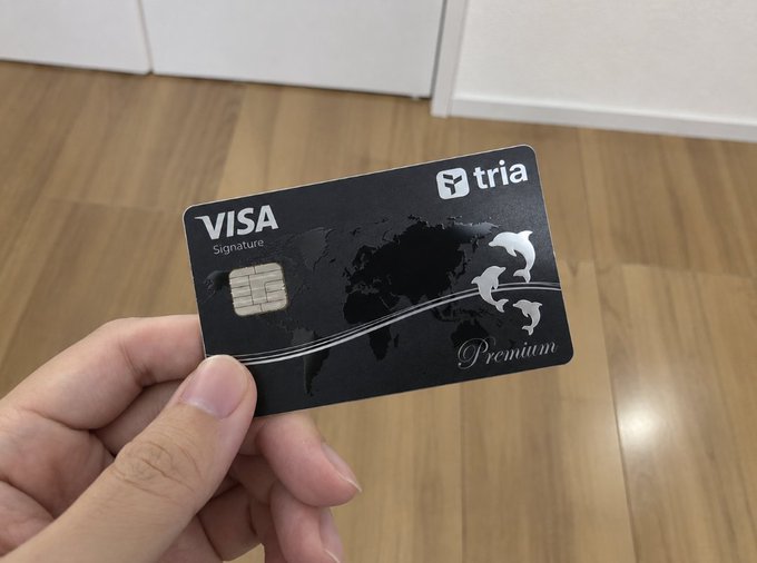 A hand holds a black Tria Visa Signature Premium credit card featuring a world map design, photographed over a wooden floor.