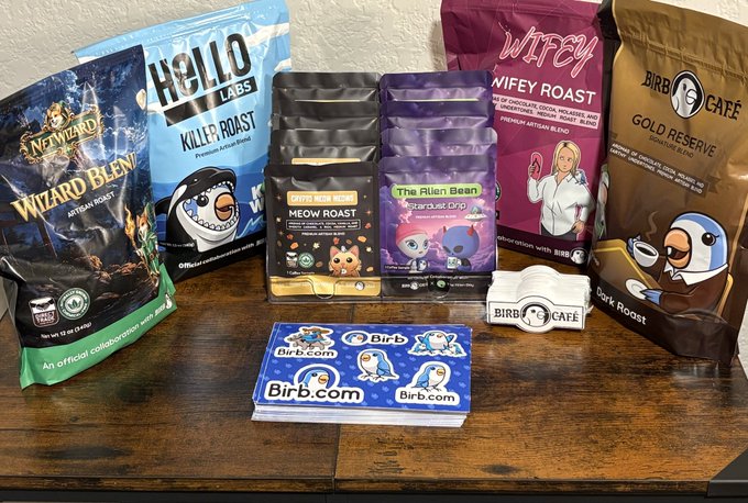 A wooden table displaying an assortment of novelty coffee bags from brands like Nerf Wizard, Hello Labs, Girb Noob, The Alien Bean, Wifey Roast, and Birb Café, accompanied by Birb.com business cards.