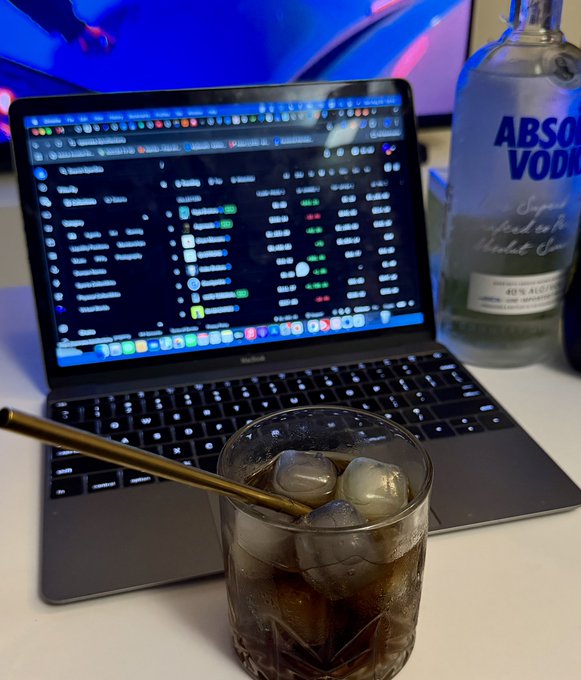 A glass of dark soda with ice and a gold straw sits in front of an open laptop displaying a financial or crypto portfolio dashboard, with a bottle of Absolut Vodka visible in the background.