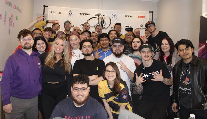 A group photo of smiling attendees at what appears to be a gaming or crypto event, standing in front of a 'SUNDIAL' and 'HYPE' branded banner, with one person in the center holding up a championship belt.
