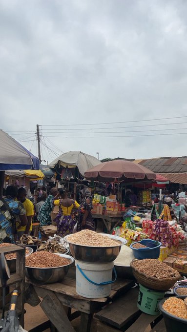 A bustling outdoor market scene in what appears to be West Africa, with vendors selling grains and goods from large bowls, baskets, and buckets under umbrellas on a cloudy day.