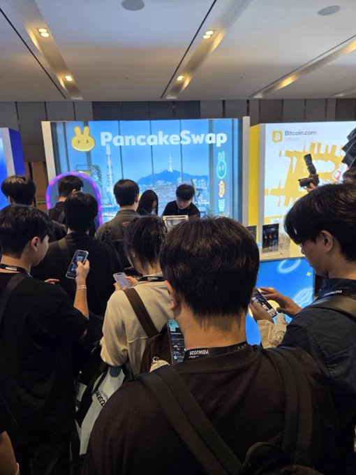 A crowd of attendees gathers at the PancakeSwap booth during KBW2026 Day 1, with many on their phones at the gacha station while the "PancakeSwap" and "Bitcoin.com" branding shines bright on the displays behind them, capturing the busy, merch-filled atmosphere described in the post.