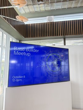 A large screen in a modern room displaying "Base Builder Meetup" with the date "October 8" and time "12-2pm" against a blue tech-themed background.