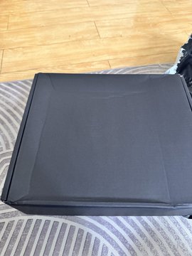 A black square cushion or seat pad sitting on a grey striped rug over a wooden floor.