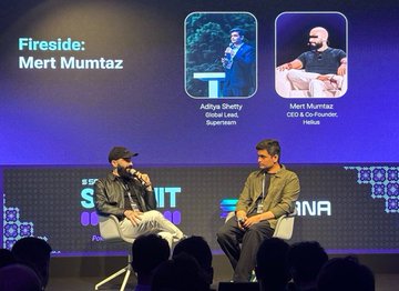 The image shows a stage with two men sitting behind a podium, likely participating in an event or panel discussion. The background features a large screen displaying the names of the speakers, including "Fireside", "Murt Muntz", and "Global Lead". There are also other people present on the stage, possibly attendees or organizers of the event.