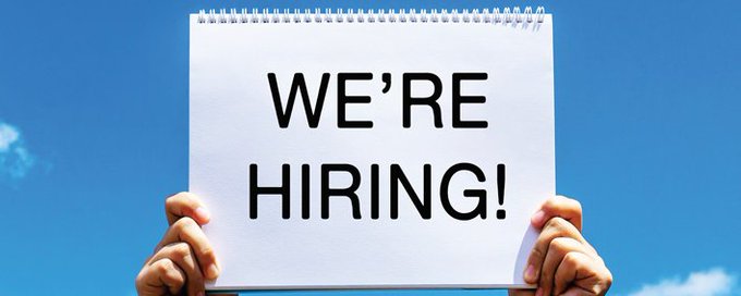 The image features a person holding up a sign that says "We're hiring!" against a blue sky background. This message seems to be promoting job opportunities or encouraging people to apply for positions at a company, likely related to the cryptocurrency industry. The person's hands are positioned in front of the sign, emphasizing its importance and visibility.