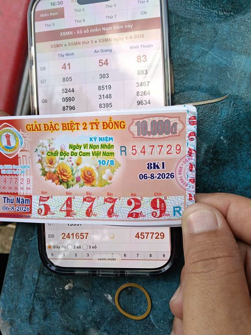 A hand holds a Vietnamese lottery ticket dated 06-8-2026 showing the winning number 547729, displayed in front of a smartphone showing lottery results.