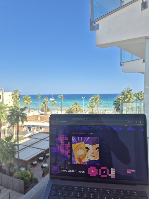 A laptop displaying a colorful NFT graphic labeled 'CHOG WORLD ORDER' sitting on a balcony overlooking a tropical beach with palm trees and turquoise water.