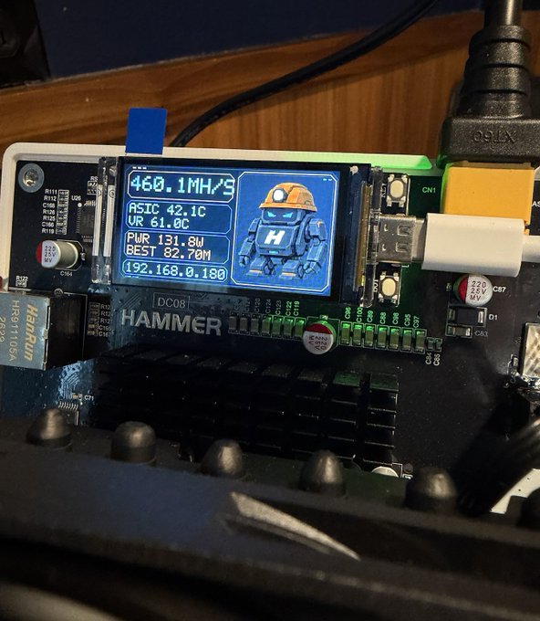 A close-up photo of an electronic device called "HAMMER" with a small blue screen displaying mining statistics such as "468.1MH/S", "ASIC 42.1C", and "PWR 131.8W", along with a graphic of a robot.