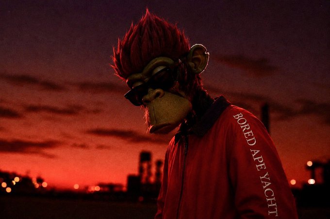A stylized 3D render of a monkey character with red spiky hair and sunglasses wearing a red 'BORED APE YACHT' jacket against a sunset cityscape.