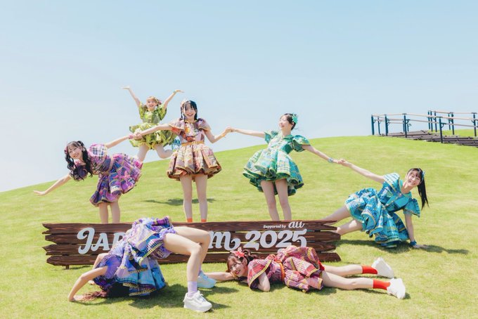 Seven young women in colorful, tropical-patterned dresses pose playfully on a grassy hill around a wooden sign reading "Japan7iem 2025" with "Supported by au" beside it. The cheerful, summery scene contrasts humorously with the caption's grumpy "My current feelings," as the group's bright outfits and lively poses capture the opposite of a bad mood.