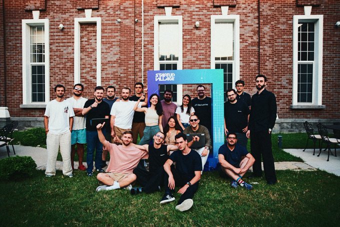 Group photo of a startup team posed outdoors on a lawn in front of a red brick building with a 'Startup AI Camp' branded sign, featuring around 15 casually dressed members.