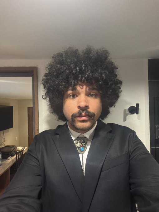 A man wearing a curly afro wig, a black suit, and a turquoise bolo tie takes a selfie in what appears to be a hotel room.