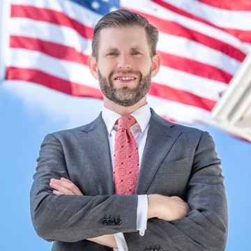 A confident-looking man in a gray suit with a pink tie stands with arms crossed in front of an American flag backdrop. The image appears to be a professional headshot related to Eric Trump, whose reported $600M+ stake in American Bitcoin has reportedly been devastated by the stock's 95%+ collapse from its peak.