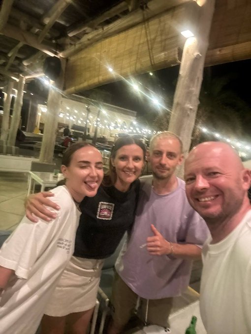 A group of four people, two women and two men, smile for a selfie at night in what appears to be an outdoor restaurant or beach club setting.