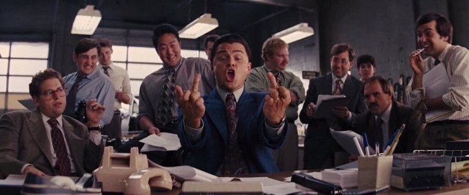 A still from the movie 'The Wolf of Wall Street' showing Leonardo DiCaprio in a blue suit making obscene gestures while surrounded by cheering office colleagues in 1990s attire.