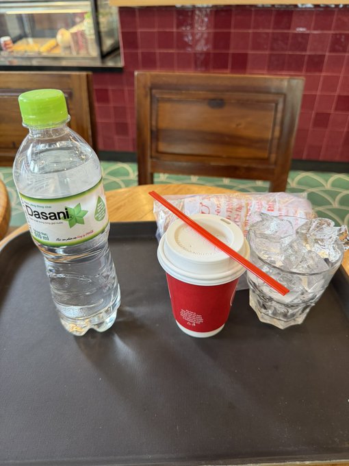 A black tray holds a Dasani water bottle, a hot drink in a red paper cup with a straw, and a glass of ice water on a wooden table in a cafe.