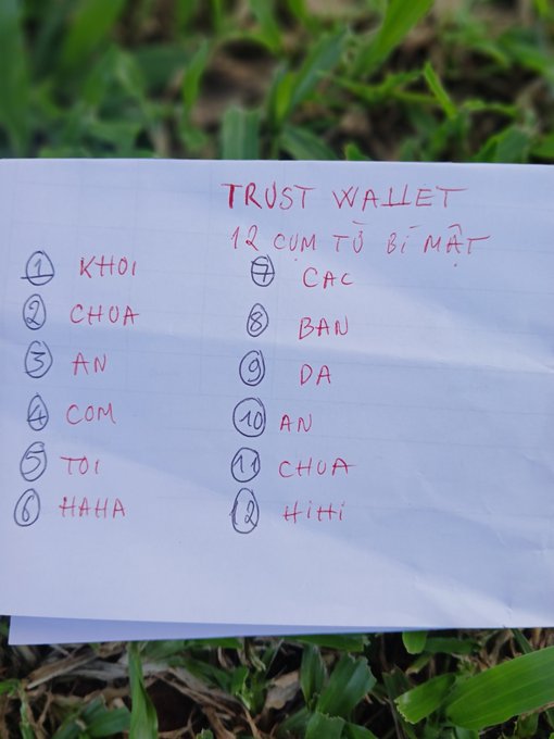 A handwritten note on paper titled "TRUST WALLET 12 CỤM TỪ BÍ MẬT" listing 12 secret recovery words in Vietnamese including KHOI, CHOA, AN, COM, TOI, HAHA, CAC, BAN, DA, AN, CHUA, and Hihi.