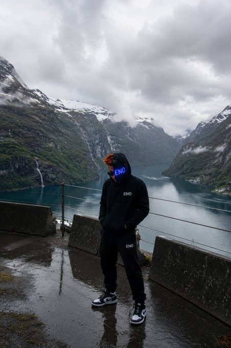 A person wearing a black 'ENO' hoodie and pants stands in front of a dramatic fjord landscape with mountains and water under a cloudy sky.