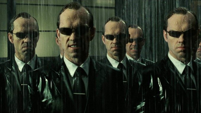 A cinematic still from The Matrix shows multiple identical versions of Agent Smith, played by Hugo Weaving, wearing black suits, white shirts, dark ties, and signature sunglasses while standing in the rain.