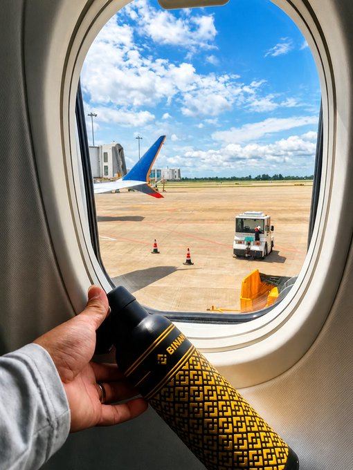 A hand holds a black and gold Binance-branded travel tumbler up to an airplane window, revealing a tail fin with blue and orange accents, ground service equipment, and traffic cones on the tarmac under a partly cloudy sky—tying into the post's playful "Binance Airlines" theme.