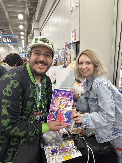 Two smiling people at a convention holding a copy of 'VEERFIENDS' comic, surrounded by merchandise in a convention hall setting.