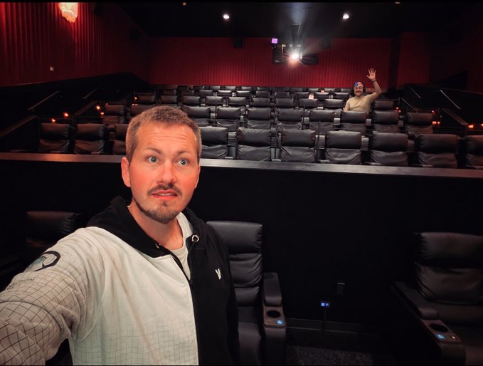 A man taking a selfie in an empty movie theater with black reclining seats and red walls, while another person in the background waves from the upper seating section.