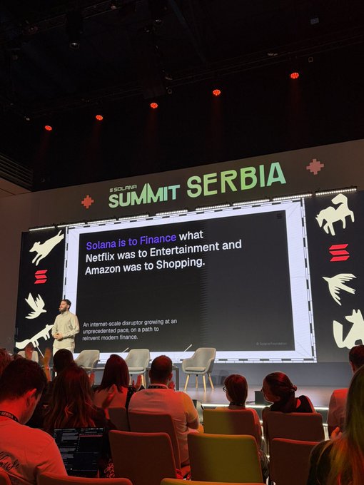 A speaker presents on stage at 'SOLANA SUMMIT SERBIA' in front of a screen reading 'Solana is to Finance what Netflix was to Entertainment and Amazon was to Shopping,' with a seated audience watching.