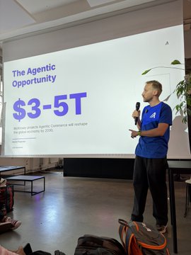 The image shows a man standing behind a projector screen, giving a presentation with a microphone. The background of the room features a white wall and a plant, creating an organized workspace for the event. There are also several bags and suitcases scattered around the room, suggesting that attendees have brought their belongings to the meeting. A laptop can be seen on one side of the image, possibly used by the presenter or other participants during the presentation. The post includes information about an upcoming workshop where attendees will learn about Algoland's cryptocurrency, specifically the AlgoXpert wallet.