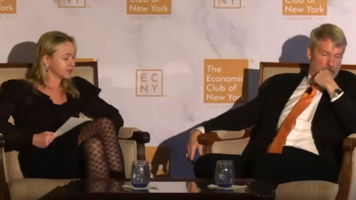 The image shows a man sitting in a chair with his legs crossed, while a woman sits next to him. They are both looking at something off-camera. The background features a white wall with a banner that reads "ECN New York". There is also a table and two cups placed on it.
