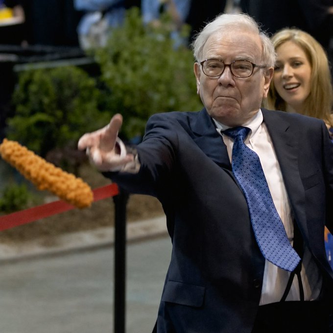 Warren Buffett, wearing a dark suit and blue tie, points forward with a displeased expression at a dirt-covered object resembling a corn dog on a red ribbon.