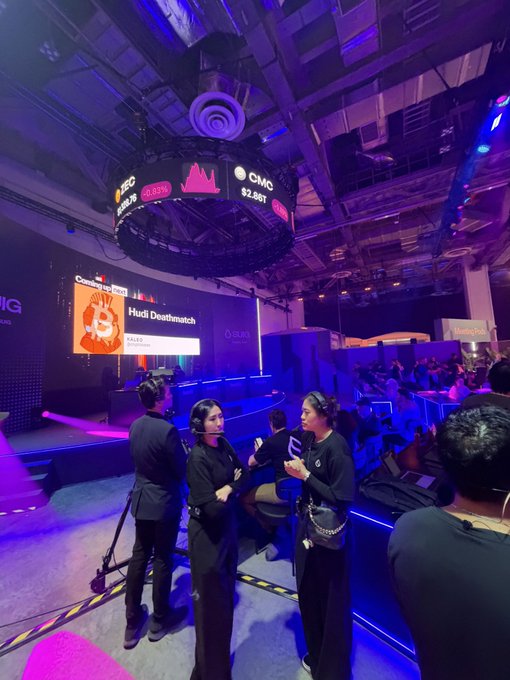 Two event staff members wearing headsets converse near a gaming arena stage, where a large screen displays "Hud Deathmatch" alongside the CMC ticker ($2.86T) and a 48.13% figure—setting the scene for an upcoming competitive esports showdown.