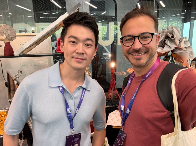 Two men smiling at the camera in an office or event space, both wearing lanyards, with a miniature Eiffel Tower visible in the background.