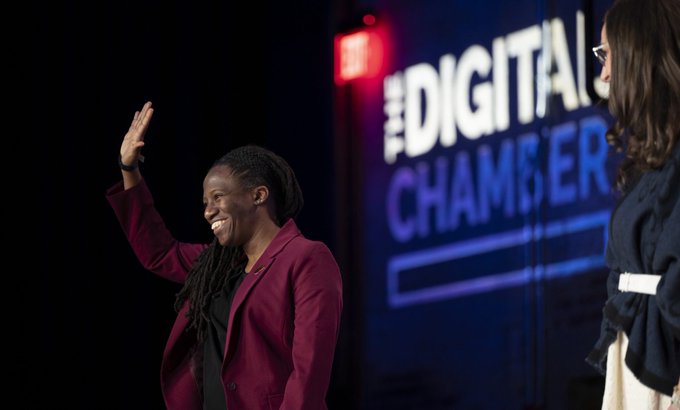 A smiling woman in a maroon blazer waves enthusiastically from a stage at the "THE DIGITAL CHAMBERS" event, representing the WVblockchain State Network Microgrant winner as highlighted in the announcement. Another person partially enters the frame from the right, suggesting a shared moment of recognition during the policy briefing.